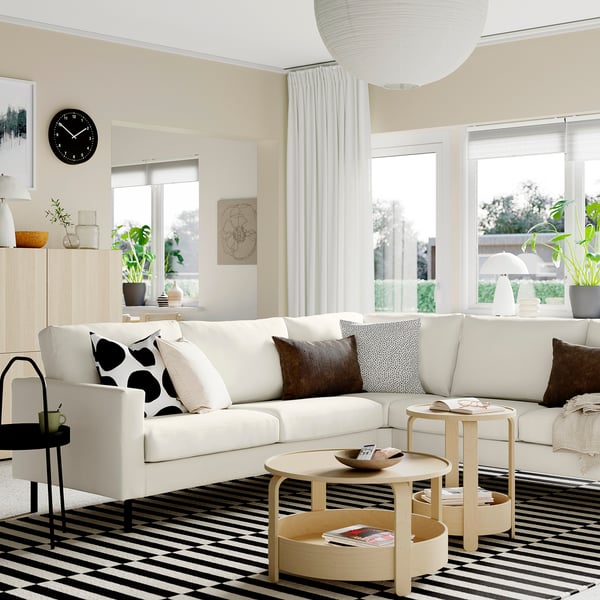 Modernes Wohnzimmer mit weißem Sofa, schwarzen und weißen Kissen, gestreiftem Teppich, Holztischen, Pflanzen und großen Fenstern.