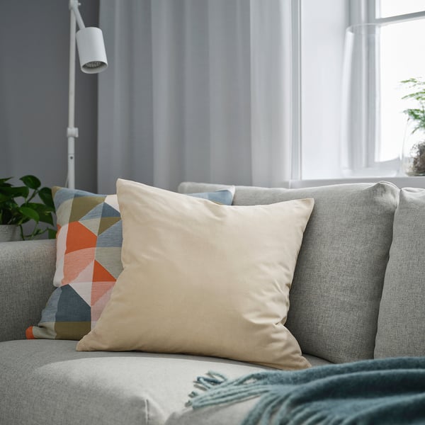 Wohnzimmer mit SANELA beigem Kissen, mehrfarbigem Kissen, grünem Überwurf, Pflanze, Lampe und Fenster mit Vorhang.