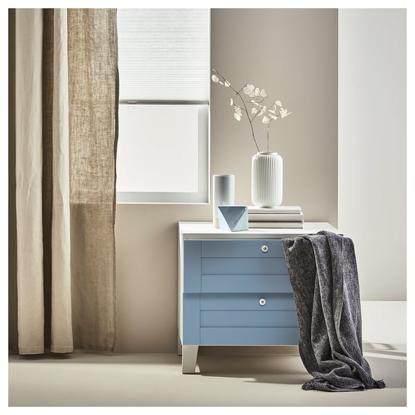 Blaue Kommode SANNIDAL mit grauen Akzenten, Decke, Vase mit weißen Blumen, drei Schubladen, beige Vorhänge.
