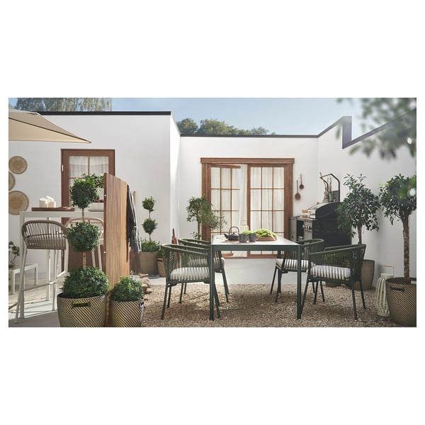 Modernes Esszimmer im Freien, weißer Haushintergrund. Schwarze Metallstühle, rechteckiger Glastisch, Topfpflanzen, Terrassendekoration.