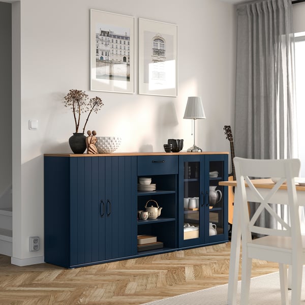 Modernes Esszimmer mit blauem SKRUVBY Sideboard, Glastüren und Porzellan. Graue Vorhänge, weiße Drucke, Holzboden.