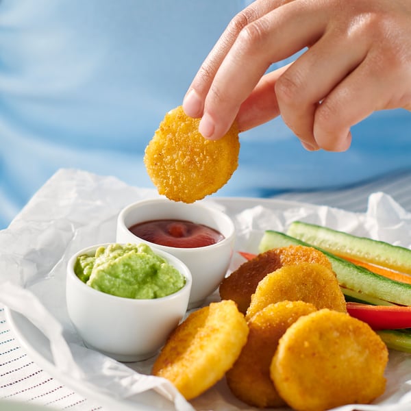 Die Hand hält ein goldbraun paniertes Weizenstück über ein Tablett mit weiteren Stücken, Gemüsesticks und zwei Dip-Saucen.