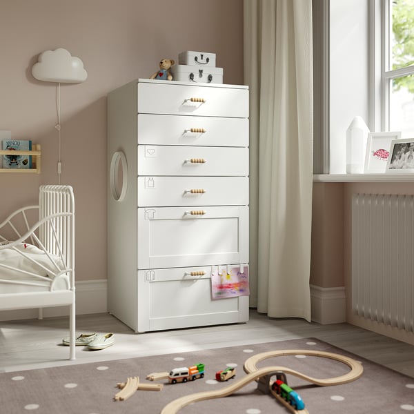 Kinderzimmer mit weißer SMÅSTAD-Kommode, 7 Schubladen, Holzgriffen. In der Nähe eines weißen Bettes, eines Fensters mit beigen Vorhängen, Spielzeug und eines Teppichs mit Tupfen.