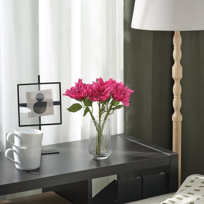 Gemütliche Schreibtischszene: leuchtend rosa SMYCKA Dahlien in klarer Vase, weiße Tassen, moderne Lampe, minimalistische Kunstwerke. Blumen ragen zwischen einfachem Dekor hervor.