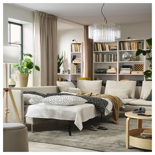 Modernes Wohnzimmer mit beigem Sofabett, weißen Kissen, schwarzer und weißer Decke, Holztisch, großem Fenster, Bücherregalen, Pflanzen und Kronleuchter.
