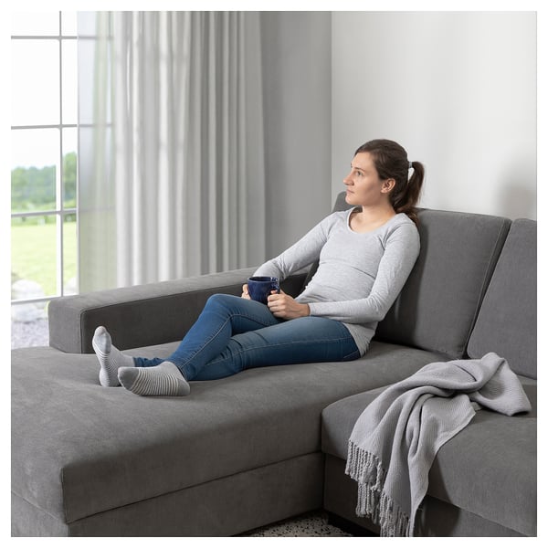 Frau entspannt sich auf grauem SÖRVALLEN-Sofa mit Kaffee in der Hand, mit großem Fenster und Vorhang dahinter.