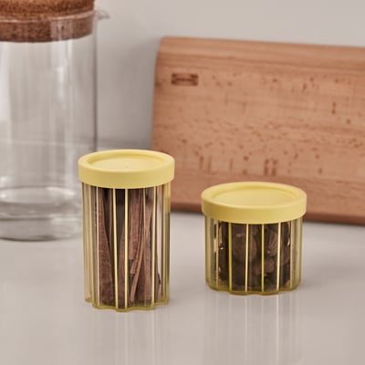 Zwei gelb-deckelnde, stapelbare Glasgefäße auf einer weißen Oberfläche mit Gewürzen, im Hintergrund eine klare Kanne und eine Holzkiste.