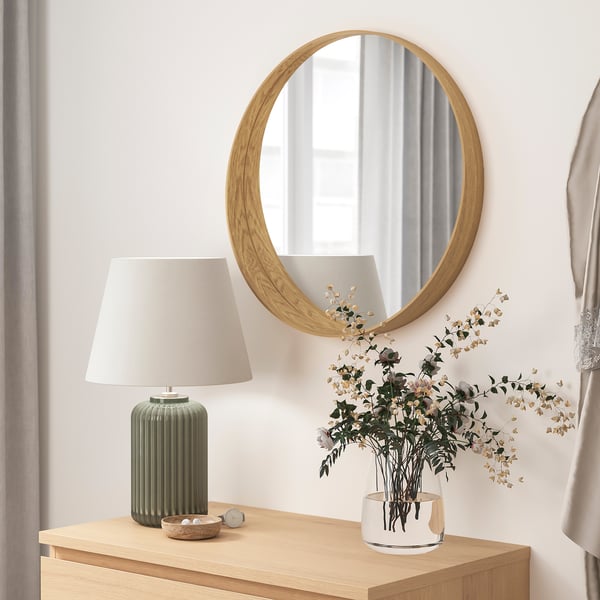 Minimalistische Raumdekoration: runder Holzspiegel, Lampe und weiße Vase mit getrockneten Blumen auf Holztisch.