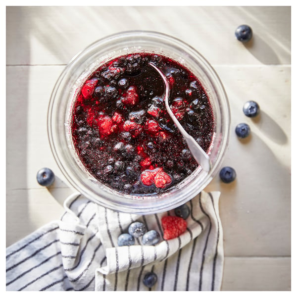 Ein durchsichtiges Glas mit gemischter Beerenmarmelade mit einem Löffel, auf ein gestreiftes Tuch gelegt, umgeben von frischen Blaubeeren und Himbeeren.