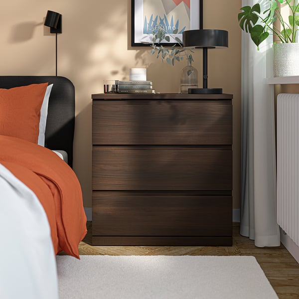 Nachttischtruhe STORKLINTA aus dunklem Holz; Enthält Schubladen, Lampe, Bücher und Vase. Neben dem Bett positioniert, kleiner als das Bett.