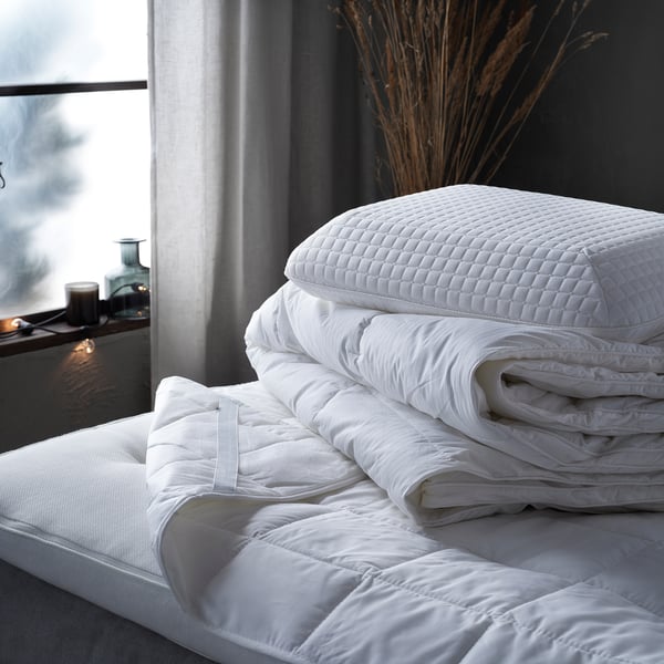 Eine kuschelige weiße Bettdecke auf einem Bett mit weichen, flauschigen weißen Kissen und gefalteten Decken mit einem Fenster im Hintergrund.