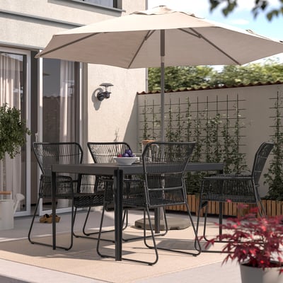 Sitzgelegenheiten auf der Terrasse mit schwarzen Stühlen von TALLSKÄR, beigem Sonnenschirm. Rattan-Design, unter einem großen Sonnenschirm, umgeben von Pflanzen.