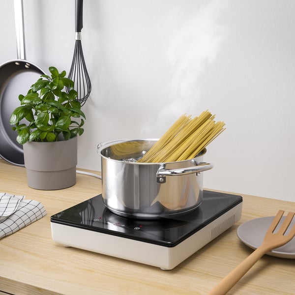 Kleine Kücheneinrichtung mit tragbarem Induktionskochfeld TILREDA, das Spaghetti kocht. Inklusive Bedienfeld, Stecker und Pflanzendekoration.