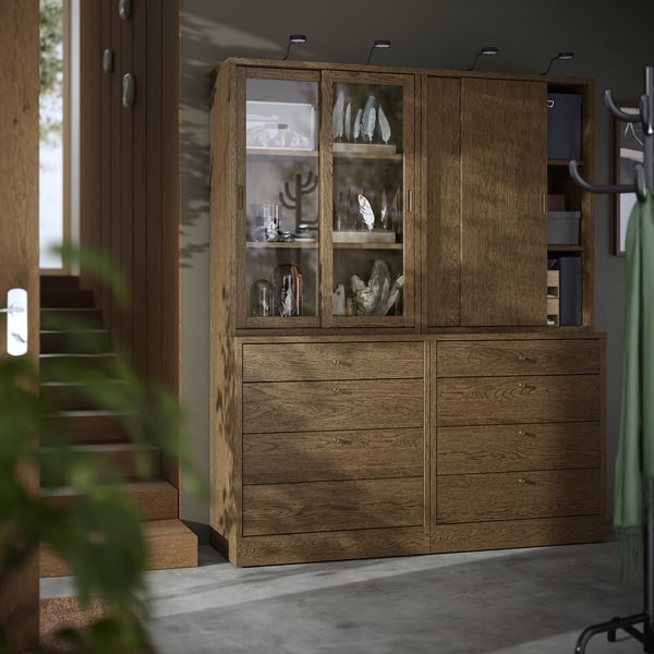 Brauner TONSTAD-Holzschrank mit Glastüren beherbergt Dekor, sitzt zwischen hohen Pflanzen und Treppen.