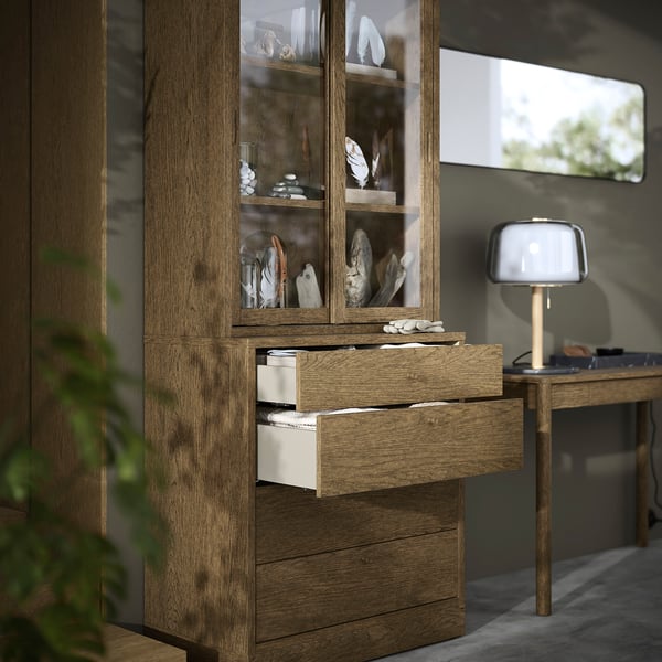 Holzschrank mit Glastüren, in denen Dekorationsgegenstände ausgestellt sind. Zwei untere Schubladen sind offen und geben den Blick frei auf organisierten Stauraum.