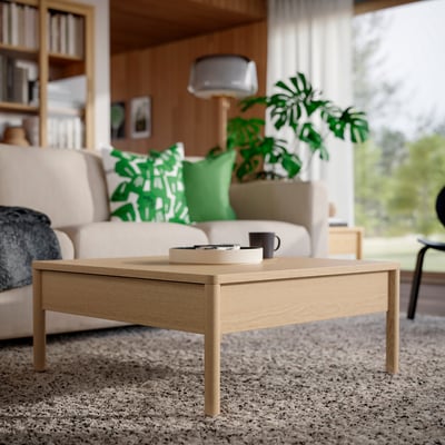 Couchtisch aus braunem Eichenfurnier mit Seitenschubladen auf beigem Teppich im gemütlichen Wohnzimmer mit Sofa, Pflanze und Bücherregal.