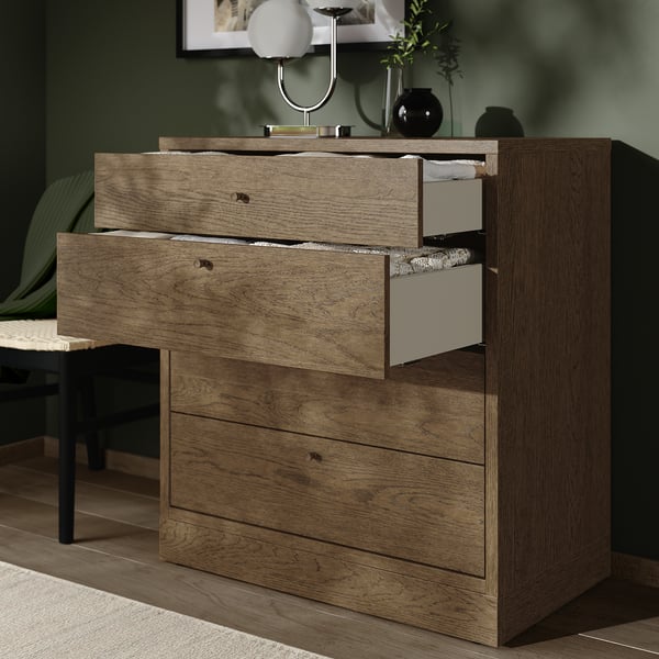 Kommode aus Holz mit 4 Schubladen, 3 offen mit Ausstellungsgegenständen im Inneren. Weiße und graue Innenschubladen, -knöpfe. Dekorative Lampe und Pflanzen obendrauf.