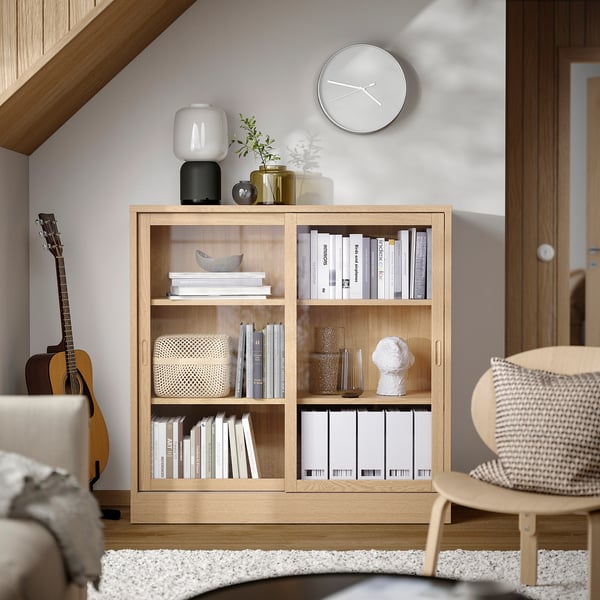 Modernes TONSTAD-Bücherregal aus heller Eiche mit Schiebetüren, in dem Bücher und Dekoration aufbewahrt werden, eingerahmt von einer weißen Uhr und Pflanzen, neben einer Gitarre.
