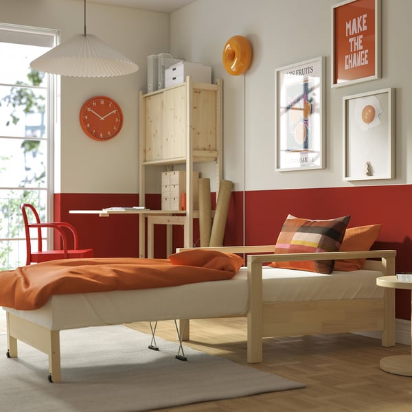 Helles Schlafzimmer mit TORNSBORG-Bett, das als Sessel zusammengeklappt ist, orangefarbener Decke, roter Uhr, Kunst und Holzschreibtisch.