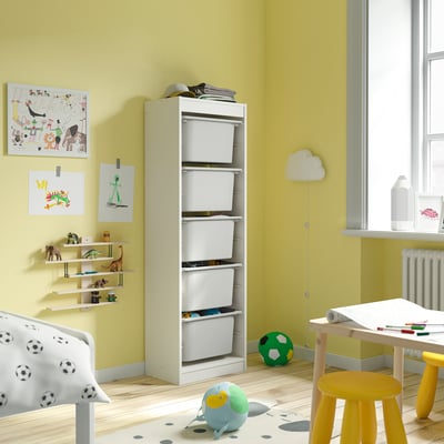 Verspieltes Kinderzimmer mit weißem TROFAST-Stauraum mit weißen Netzboxen und Plastiktabletts, das in der Nähe eines Fensters mit gelben Wänden aufgestellt ist.