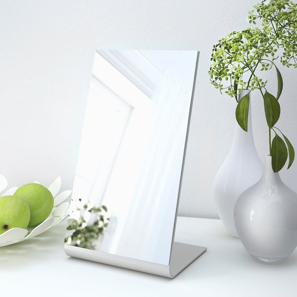 Ein rechteckiger Tischspiegel auf einem Ständer neben einer weißen Vase mit grünen Pflanzen und einer Obstschale.
