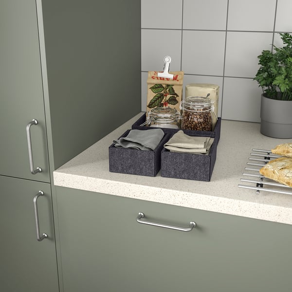 Küchentheke mit grünen Schränken und gesprenkelter Arbeitsplatte. Zwei graue Schachteln mit Stoffservietten, Gläsern und einer Papiertüte mit Kaffeebohnen und Klammern.