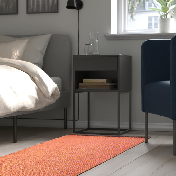 Modernes Schlafzimmer mit einem grauen Bett, einem schwarzen Nachttisch mit Büchern und einem bunten Teppich, der dem Raum Wärme und Textur verleiht.