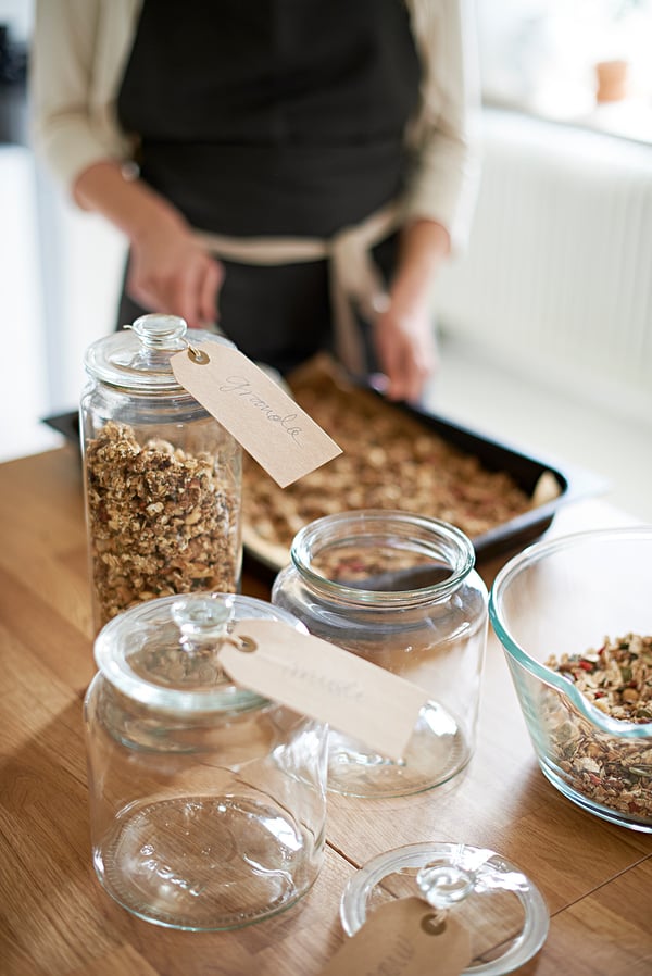 Eine Person kocht Müsli in einer Pfanne auf einer hölzernen Küchentheke mit beschrifteten Gläsern mit hausgemachtem Müsli.