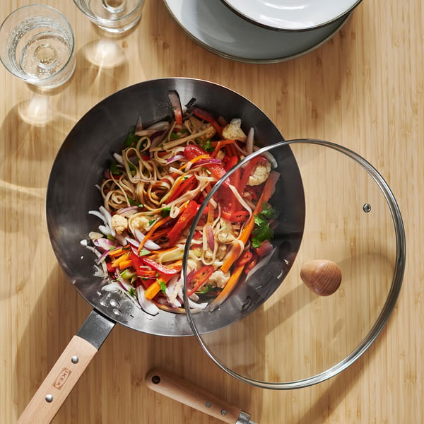 Ein Wok aus schwarzem Kohlenstoffstahl auf einem Holztisch, gefüllt mit buntem Pfannengemüse und Nudeln, steht neben einem Glasdeckel und leeren Weingläsern.