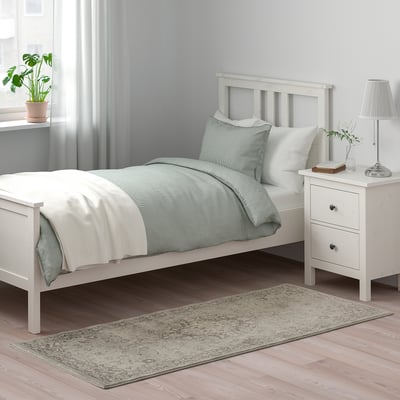 Schlafzimmer mit grauem VEDBÄK-Blumenteppich neben weißem Bett, ergänzt durch hellgraue Vorhänge.