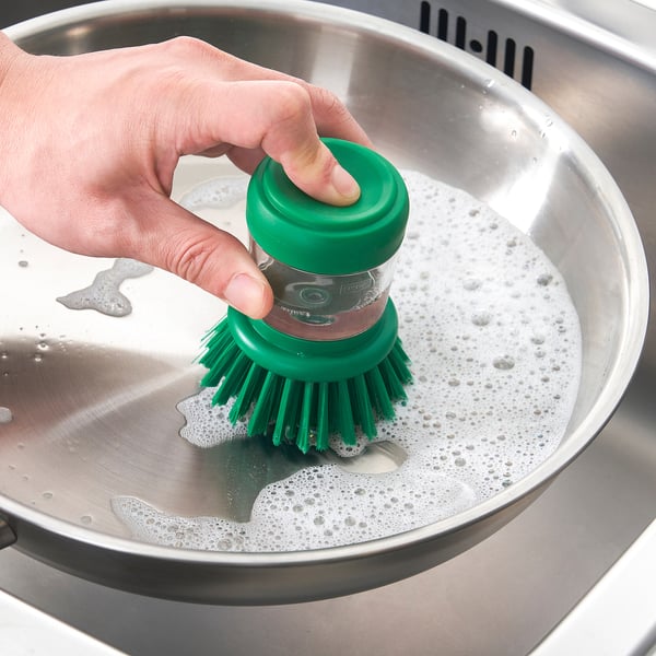 Eine Hand hält eine grüne Bürste und taucht sie in ein Edelstahlwaschbecken mit Seifenwasser, um die Fähigkeit der Bürste zu demonstrieren, Seife abzugeben.