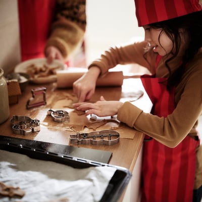 Ein Kind mit einer Schürze hilft beim Keksbacken. Die Schürze lässt sich mit einem Nackenriemen verstellen, sodass Kleidung nicht verschmutzt wird.