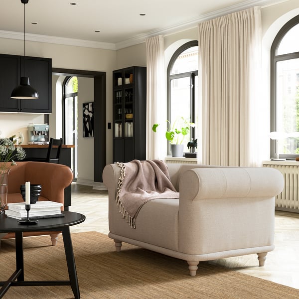 Modernes Wohnzimmer mit beigem VISKAFORS-Sessel, hölzernem Couchtisch, schwarzen Vorhängen und großen Bogenfenstern.