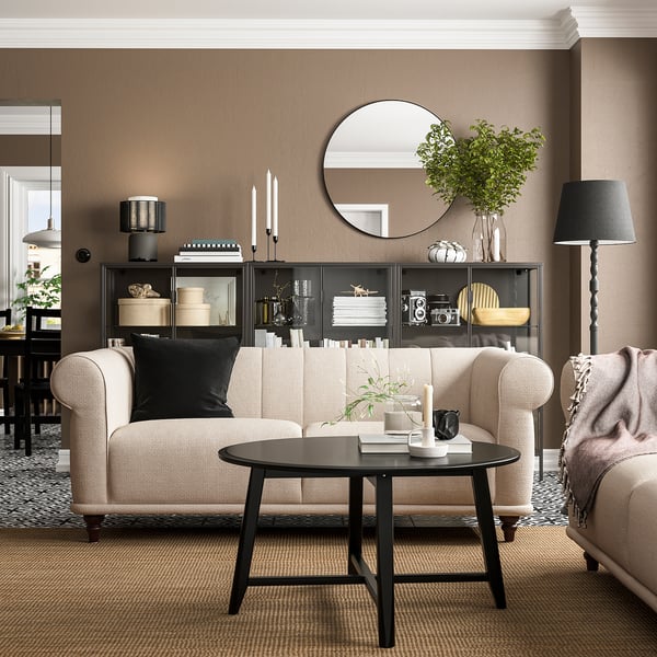 Modernes Wohnzimmer mit hellem VISKAFORS-Sofa, schwarzem Couchtisch und dunklem Holzregal mit Spiegel und Pflanzen.