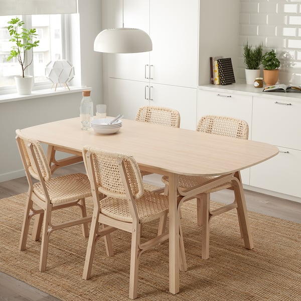 Esszimmergarnitur aus Bambus mit glattem Tisch, abgewinkelten Beinen und geflochtenen Stühlen auf Juteteppich, weiße Küche, großes Fenster.