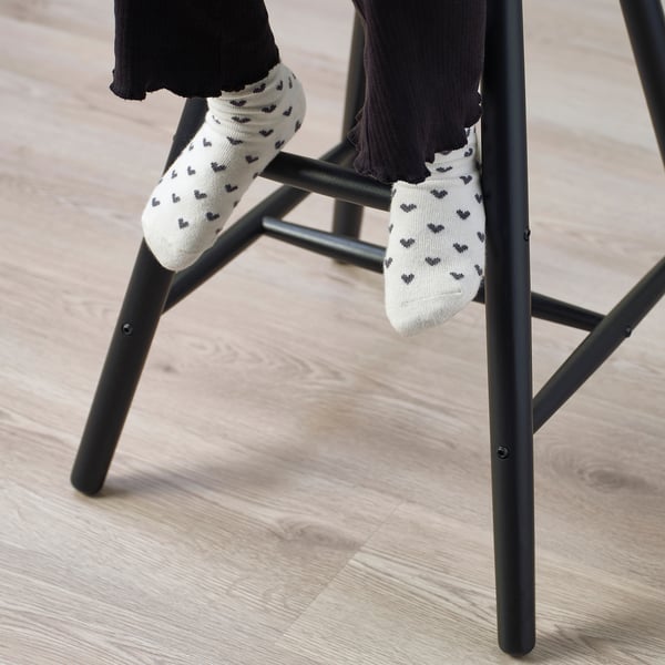 A child sits on a junior chair, feet on its bars, wearing white socks with black hearts. The chair gives proper seat height and foot support.