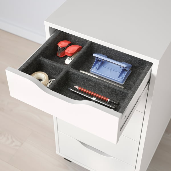 Open white ALEX desk drawer revealing organised office supplies like tape, stapler, pens, and a pencil case on grey felt liner.