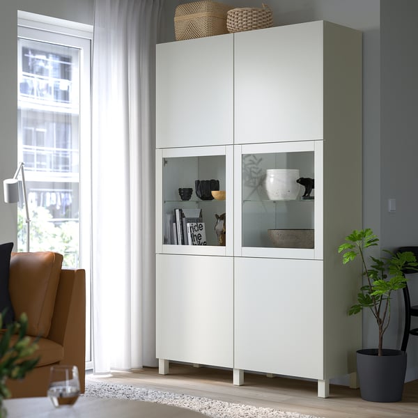 BESTÅ glass-door cabinet near window, with adjustable shelves and plant.