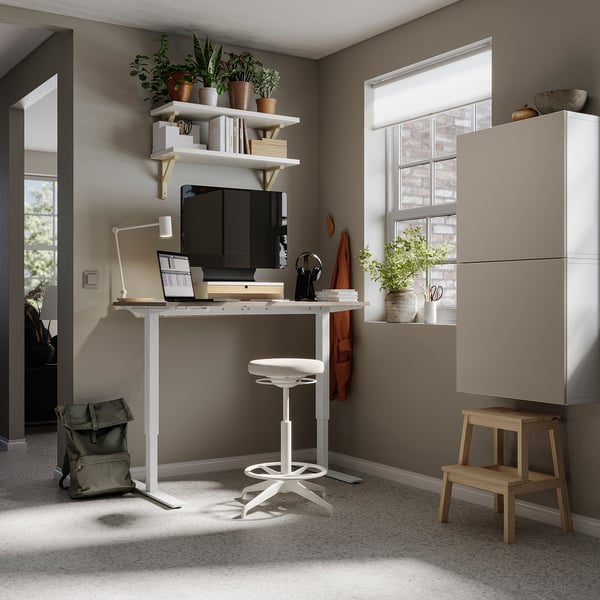 Home office with BESTÅ desk, laptop, monitor, stool, shelves with plants, and beige storage unit. Modern and organised.