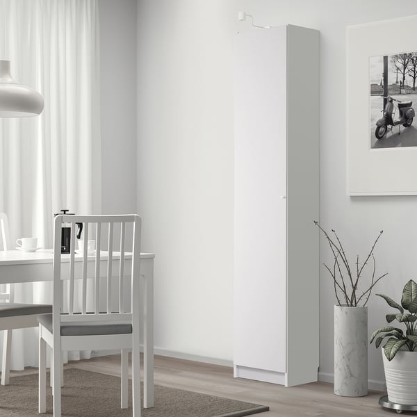 White BILLY bookcase in minimalist room, next to dining table with chairs, tall sleek design, beside large window with curtains.