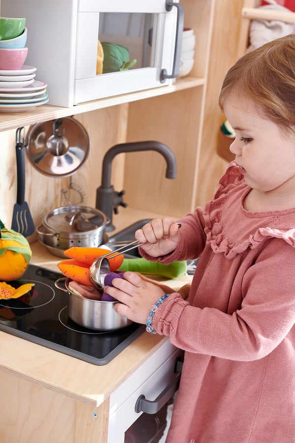 A young girl plays with DUKTIG toy kitchen, cutting orange veggies on a black stovetop, wearing a pink sweater.