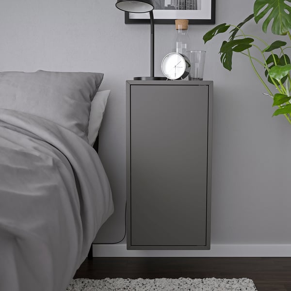 Modern grey wall-mounted cabinet, part of an ikea EKET series, holding a clock, and a lamp, mounted beside a bed.