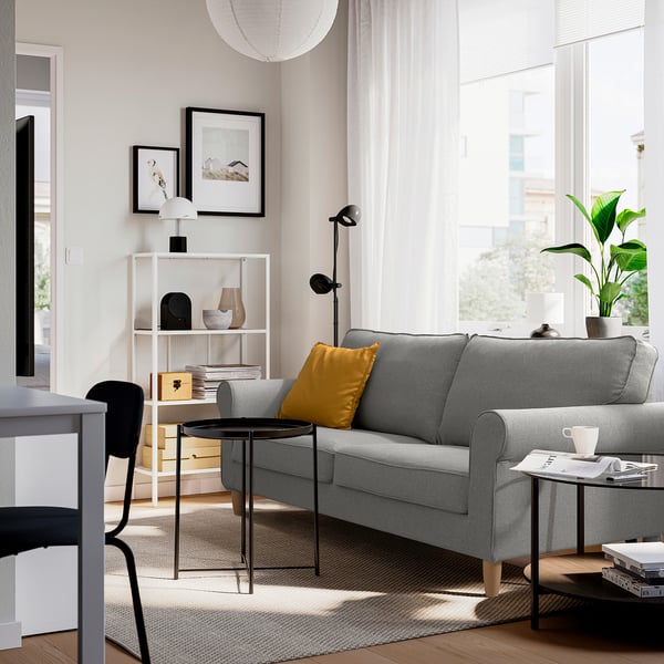 Modern living room with grey GÄLLTOFTA sofa, yellow pillow, black side table, white curtains, large window, and plants.