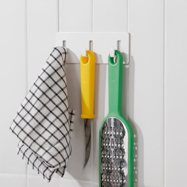 Three hooks on a white wall hold a chequered cloth, a yellow knife, and a green grater. Organises kitchen tools efficiently.