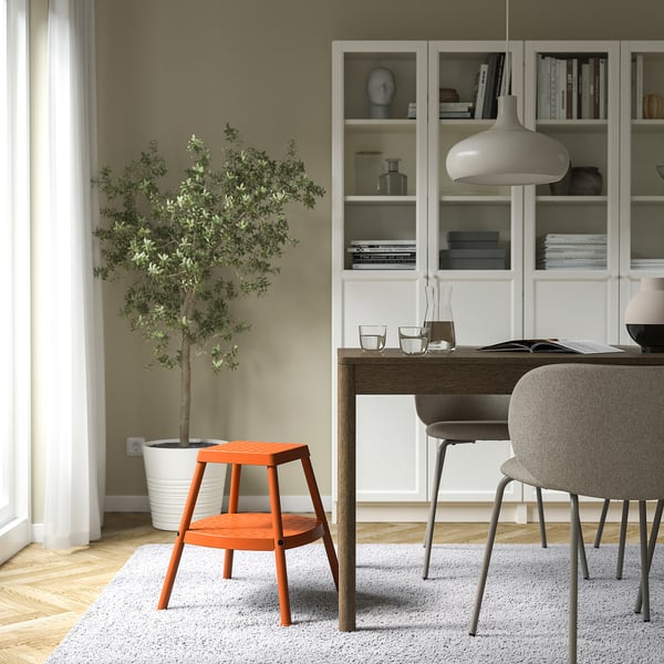 Minimalist room with orange HADDARP stool near plant, contrasting with neutral decor.