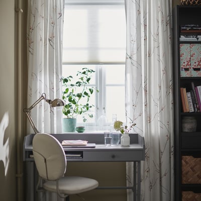 Home office setup: white desk, chair, lamp, plant, window, bookshelf.