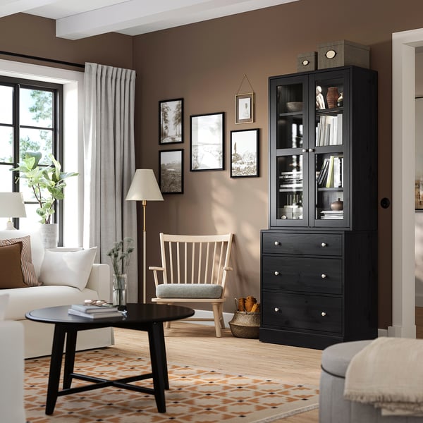 Black HAVSTA cabinet with glass doors books. Beside a white chair and black coffee table with vase. Room decorated with photos, lamp, and orange rug.