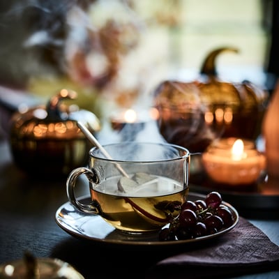 A cosy autumn table setting with a steaming cup of tea on a glass mug, grapes, and warm lighting.