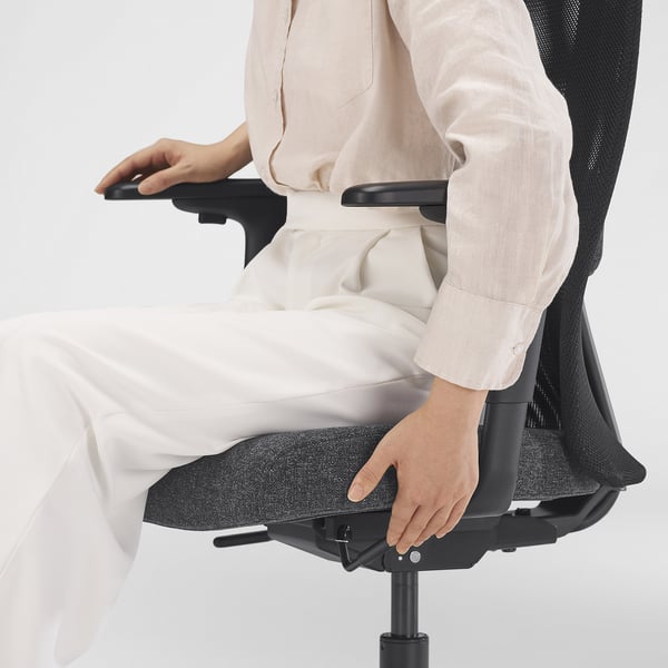 Person adjusting ergonomic office chair, targeting lumbar support. Chair has mesh back, height-adjustable seat, and tilt lock feature.