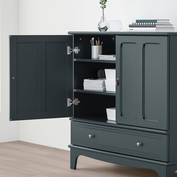 Dark green cabinet with open doors revealing shelves holding books and a jar of pens, against a white wall.